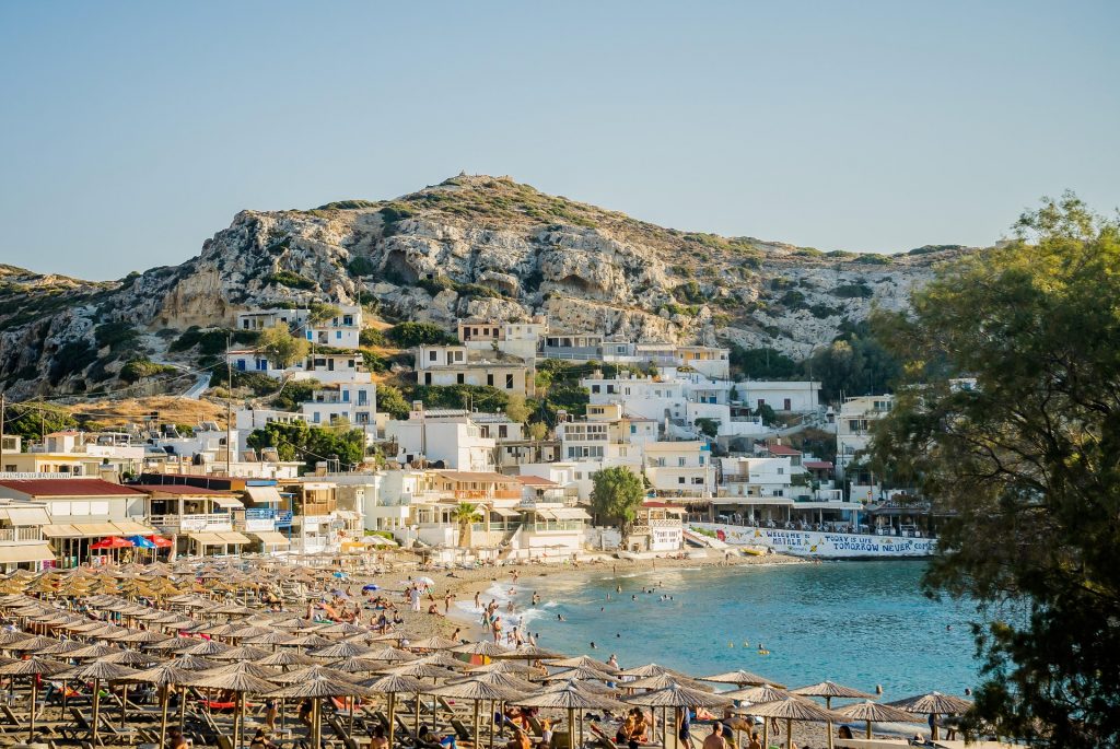 Sun-soaked Greek beach lined with straw parasols and sunbeds, with swimmers in turquoise waters and a hillside village in the background.