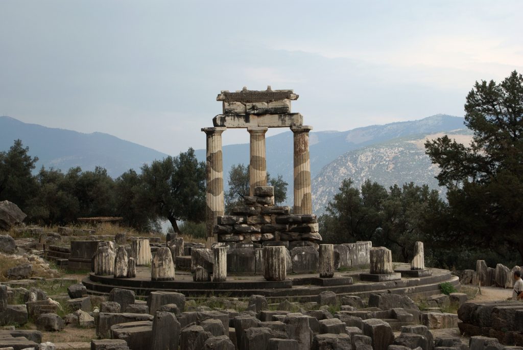 Ancient ruins of the Tholos of Delphi, with three standing marble columns framed by olive trees and distant mountains.
