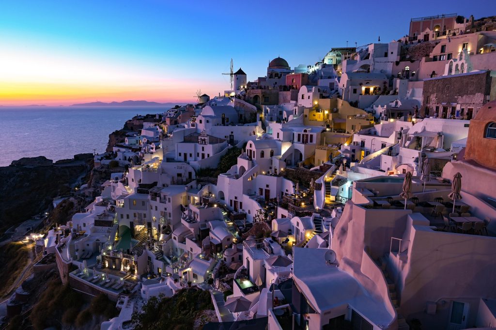 Twilight view of Oia, Santorini, with whitewashed cliffside buildings illuminated by warm lights against a vivid sunset over the Aegean Sea.